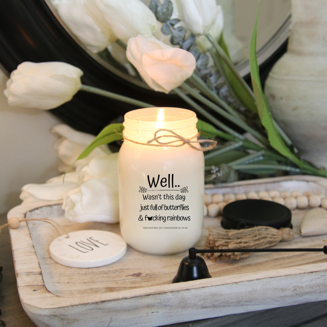 A 16 oz mason jar soy candle burning  with a label that says "Well..Wasn't this day just full of Butterflies & F*@king Rainbows."