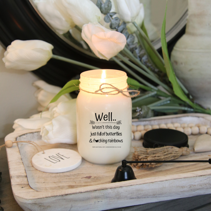 A 16 oz mason jar soy candle burning  with a label that says "Well..Wasn't this day just full of Butterflies & F*@king Rainbows."