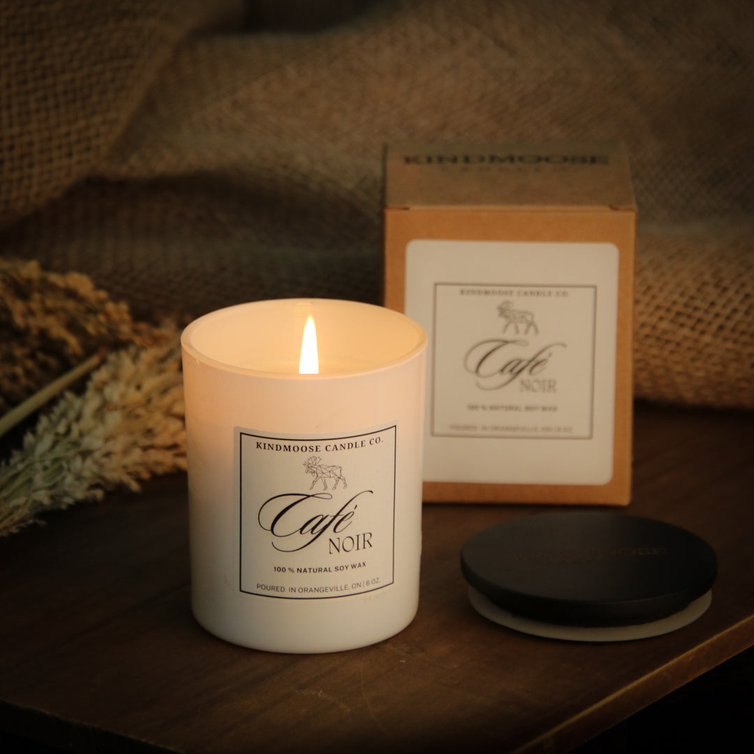 A lit candle labeled 'Café Noir' sits on a wooden surface surrounded by coffee beans. The candle is white with a simple, elegant label and is accompanied by a matching gift box and a wooden lid. The background features a warm, softly blurred ambiance, suggesting a cozy, intimate setting