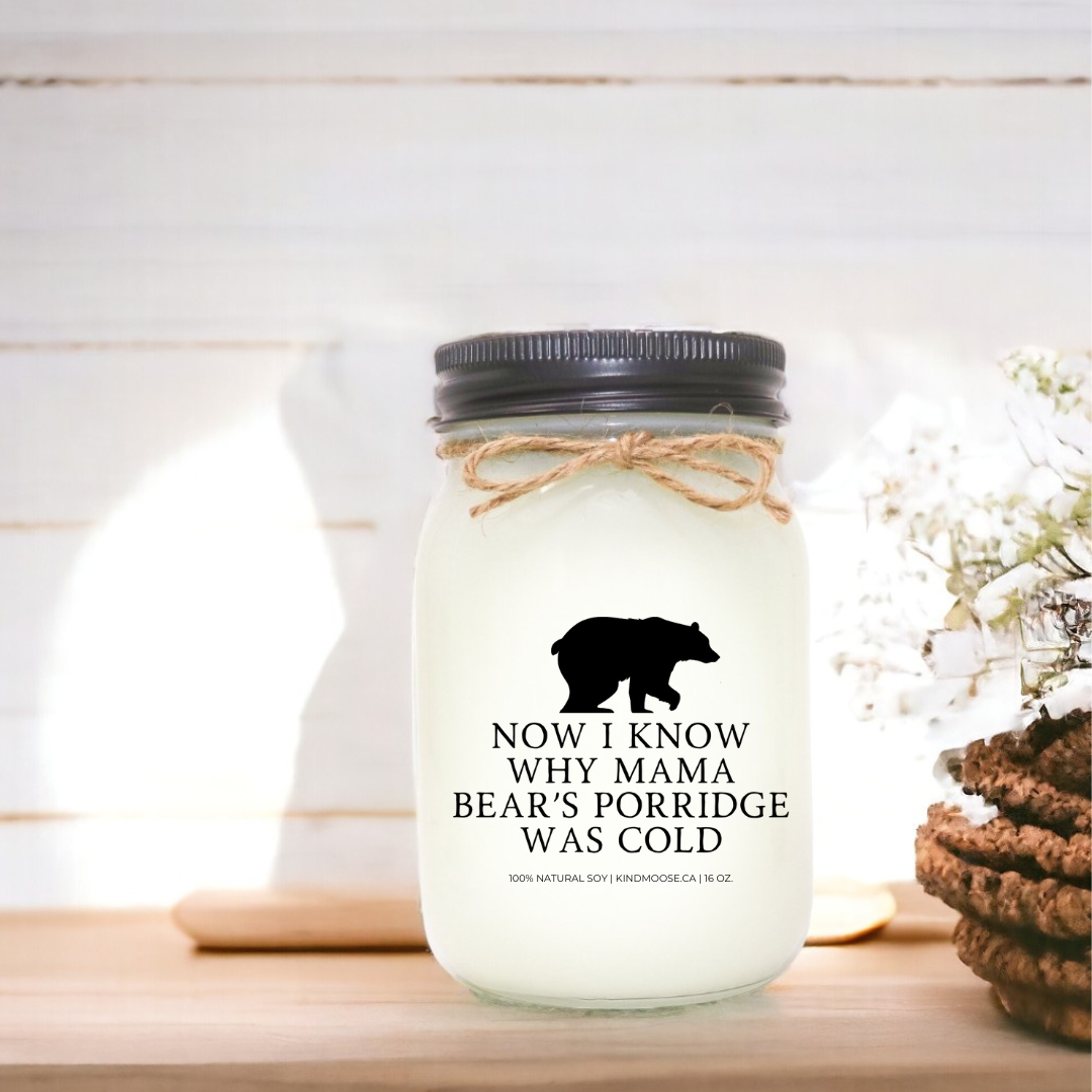 A scented candle with the label "now i know why mama bear's porridge was cold" displayed against a wooden backdrop alongside flowers and cookies.