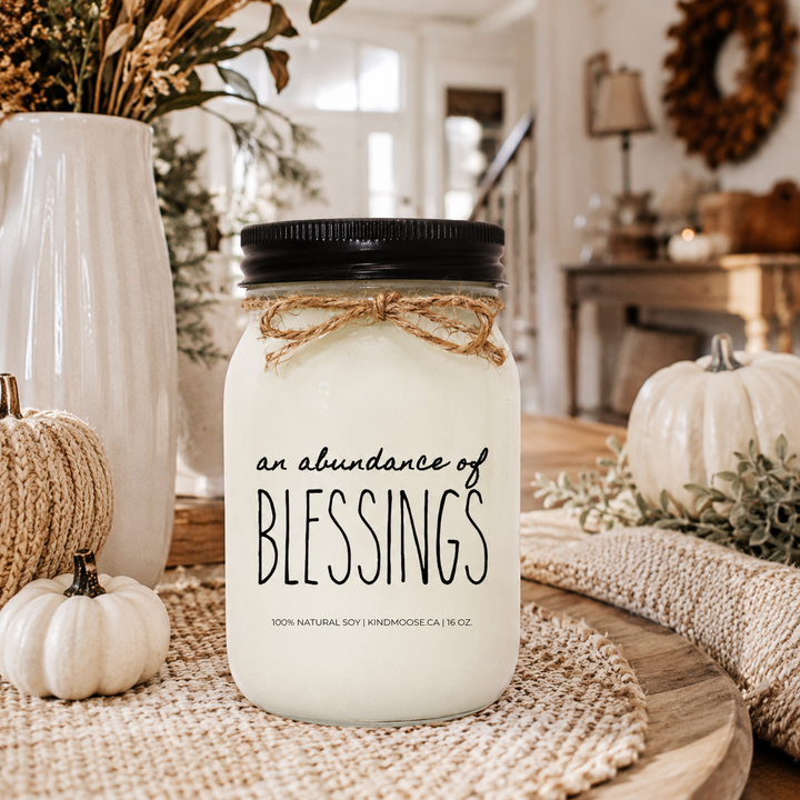 AN ABUNDANCE OF BLESSINGS soy candle with black lid displayed on an elegant harvest entry table featuring neutral pumpkins, autumn greenery, woven accents, and warm fall décor.