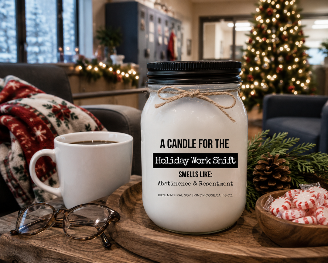A KINDMOOSE "A Candle for the Holiday Shift" 16 oz soy candle with a black lid sits on a reclaimed wood coffee table inside a cozy hospital staff lounge decorated for Christmas. A white coffee mug, reading glasses, cedar branches, pinecones, and a folded fleece blanket create a warm and comforting scene for those working through the holidays.