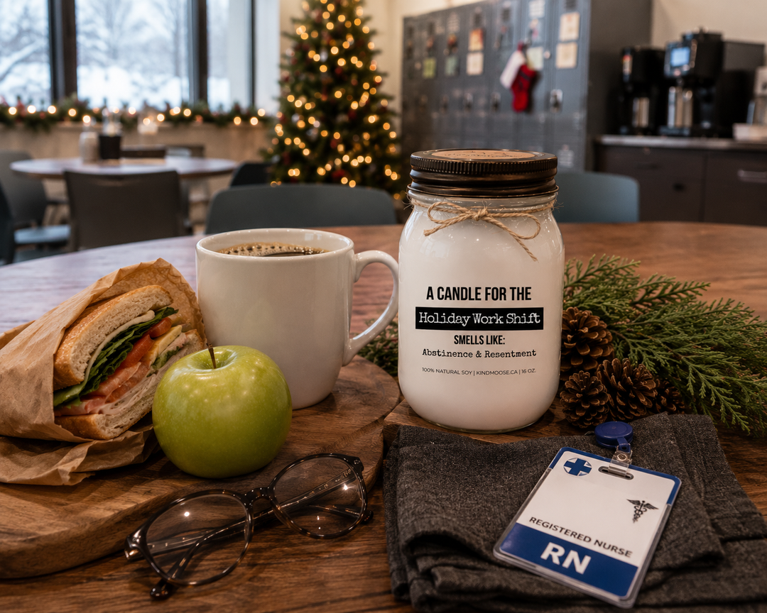 A KINDMOOSE "A Candle for the Holiday Shift" 16 oz soy candle with a distressed bronze lid rests on a reclaimed walnut breakroom table during a holiday meal break. A fresh cup of coffee, wrapped sandwich, green apple, hospital ID badge, cedar branches, and pinecones capture the reality of working Christmas shifts while everyone else celebrates.