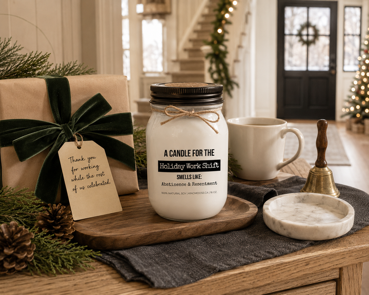 A KINDMOOSE "A Candle for the Holiday Shift" 16 oz soy candle with a distressed bronze lid is displayed on a reclaimed white oak entry console beside a beautifully wrapped Christmas gift, handwritten thank-you tag, white ceramic mug, cedar branches, pinecones, and a vintage brass bell. This thoughtful holiday gifting scene honors nurses, first responders, flight crews, and essential workers who spend Christmas caring for others.