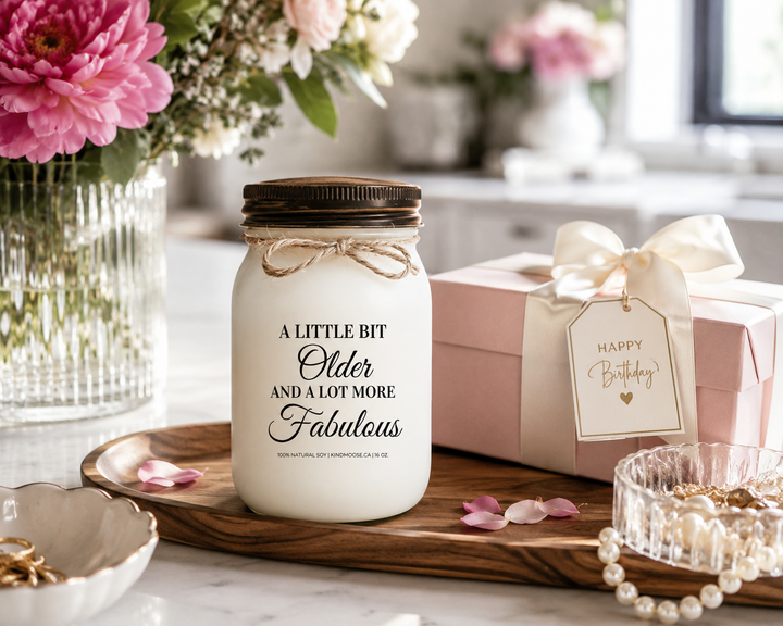 A LITTLE BIT OLDER AND A LOT MORE FABULOUS customizable soy candle with a black lid displayed on a reclaimed white oak console table in a luxury Canadian modern farmhouse. Fresh blush peonies, a gift-boxed keepsake, pinecones, and elegant birthday décor create a sophisticated birthday celebration scene filled with warmth and style.