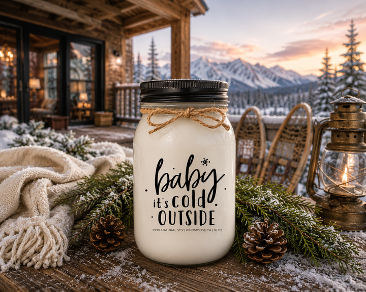 BABY IT'S COLD OUTSIDE KINDMOOSE candle with a black lid displayed on a reclaimed white oak ski chalet porch table overlooking a snow-covered mountain landscape at sunset, surrounded by fresh cedar branches, pinecones, an ivory wool scarf, a brass lantern, and vintage wooden snowshoes.