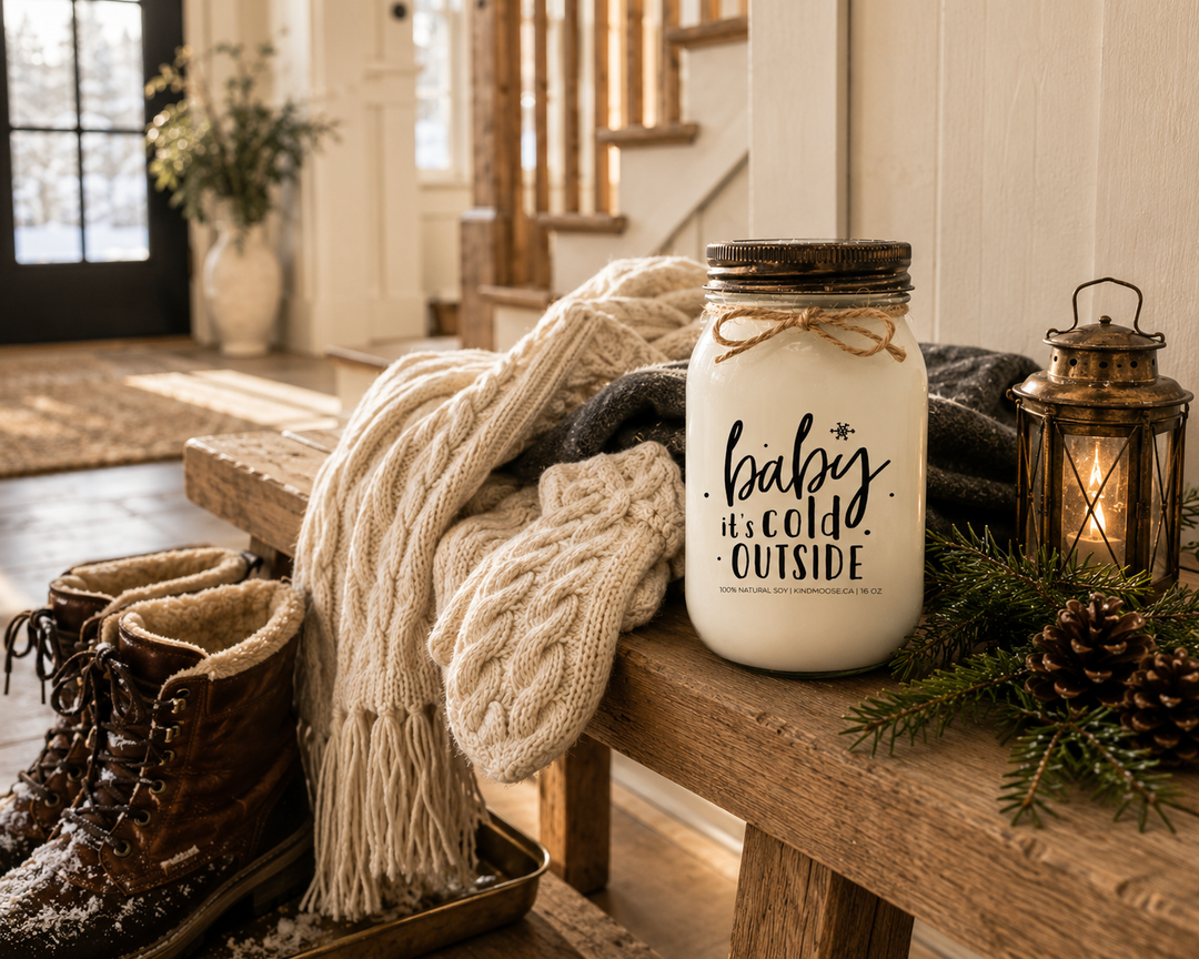 BABY IT'S COLD OUTSIDE KINDMOOSE candle with a distressed bronze lid resting on a reclaimed white oak entry bench beside snow-dusted boots, a cream knit scarf, mittens, cedar branches, pinecones, and a brass lantern inside a warm luxury farmhouse foyer after a snowy winter walk.