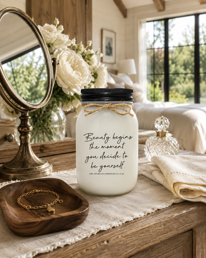 BEAUTY BEGINS THE MOMENT YOU DECIDE TO BE YOURSELF inspirational KINDMOOSE candle with a matte black lid displayed on a reclaimed white oak bedroom vanity surrounded by white garden roses, a brass mirror, crystal perfume bottle, gold bracelet, and soft linen in a bright luxury modern farmhouse bedroom.