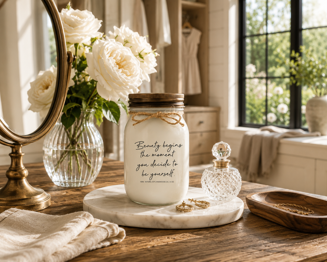BEAUTY BEGINS THE MOMENT YOU DECIDE TO BE YOURSELF KINDMOOSE candle with a distressed bronze lid on a reclaimed white oak dressing table with a marble tray, white garden roses, crystal perfume bottle, gold earrings, and antique brass mirror inside a luxury Canadian modern farmhouse dressing room filled with morning sunlight.