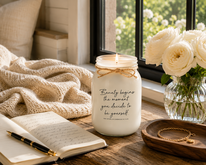 BEAUTY BEGINS THE MOMENT YOU DECIDE TO BE YOURSELF burning KINDMOOSE candle without a lid on a reclaimed white oak window seat beside an open journal, brass fountain pen, white garden roses, walnut tray, and knit throw in a peaceful luxury modern farmhouse reading nook overlooking a summer garden.