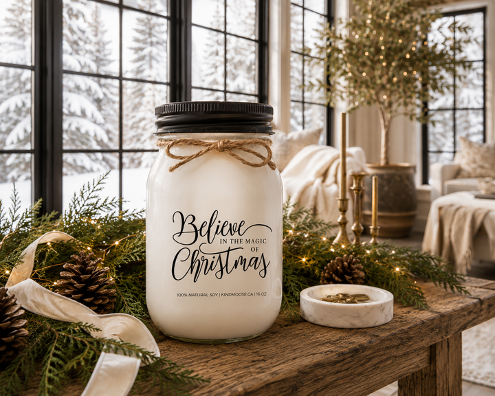 BELIEVE IN THE MAGIC OF CHRISTMAS candle displayed as an elegant Christmas gift on a reclaimed white oak library console, styled with an ivory wrapped present, cedar branches, pinecones, carved wooden angel, antique brass sleigh bell, and bright winter sunlight overlooking a snowy landscape.