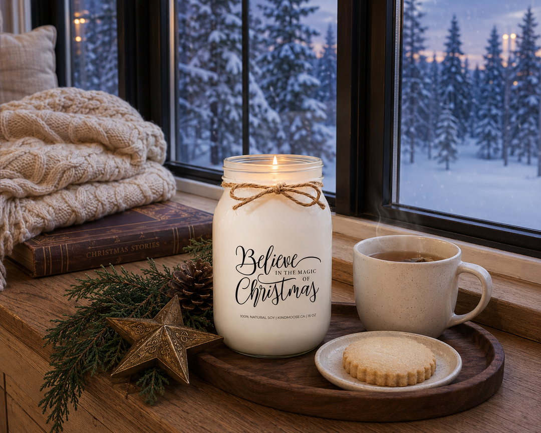 BELIEVE IN THE MAGIC OF CHRISTMAS candle burning on a reclaimed white oak window seat overlooking a peaceful snow-covered evergreen forest, styled with a cable-knit throw, herbal tea, shortbread cookie, cedar sprigs, and soft Christmas Eve lighting.