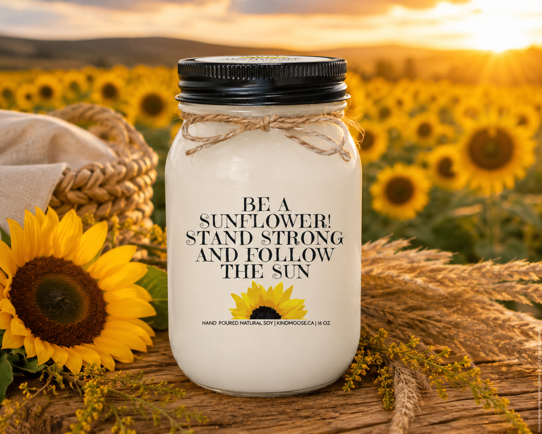 BE A SUNFLOWER! STAND STRONG AND FOLLOW THE SUN soy candle displayed on a rustic harvest table in a sunflower field at golden hour, surrounded by fresh sunflowers, warm grasses, and beautiful countryside views.