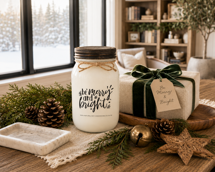 Be Merry and Bright customizable Christmas candle with a distressed bronze lid displayed on a reclaimed white oak library console beside a beautifully wrapped ivory gift with emerald velvet ribbon, fresh cedar branches, pinecones, an antique brass sleigh bell, and a carved wooden star in a luxury Canadian farmhouse library.