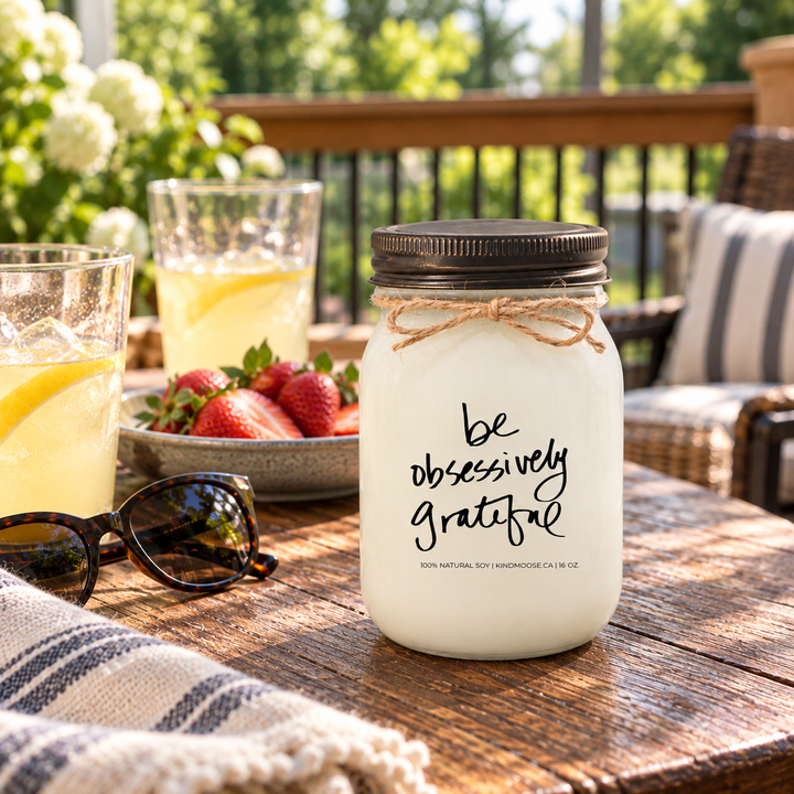 KINDMOOSE Be Obsessively Grateful candle with distressed bronze lid on a sunny backyard deck, beside lemonade and strawberries shared during an afternoon with friends.