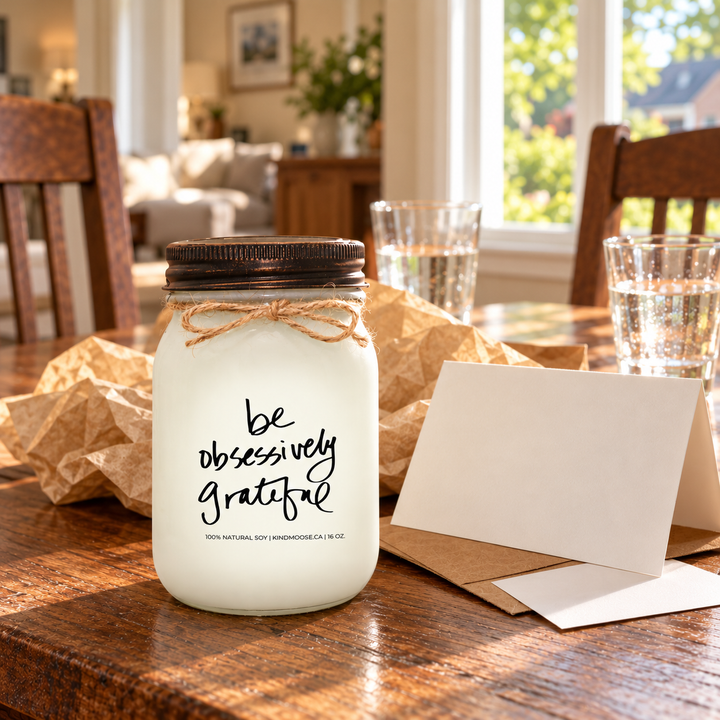 KINDMOOSE Be Obsessively Grateful 16 oz candle with distressed bronze lid presented as a thoughtful appreciation gift with a greeting card and kraft wrapping on a sunlit dining table.