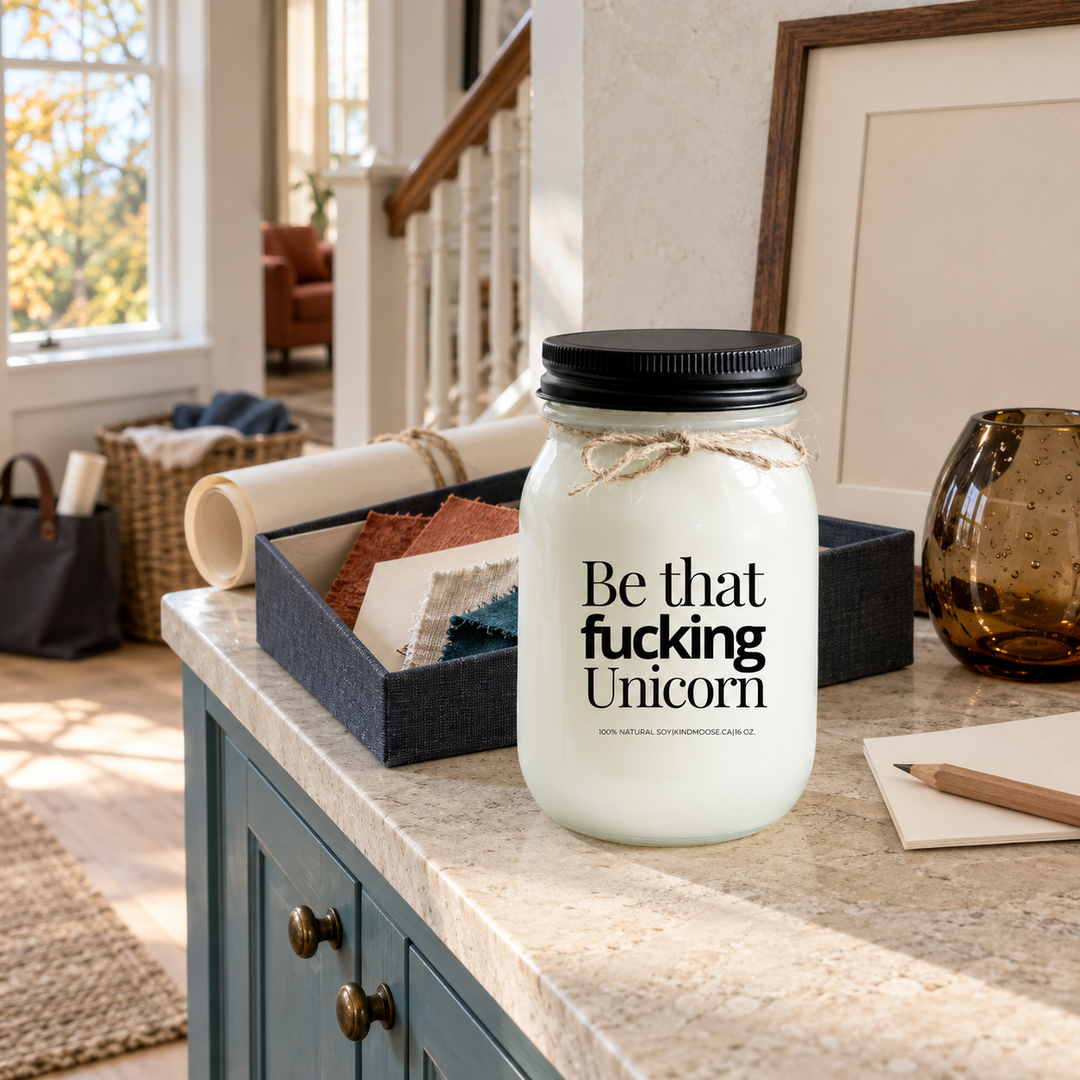 Be That Fucking Unicorn KINDMOOSE candle with black lid in a bright cottage alcove styled with creative materials and blue-green cabinetry.