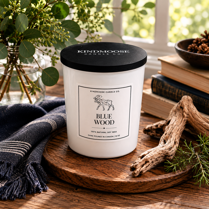 Blue Wood 6 oz soy candle by KINDMOOSE Candle Co. displayed on a rustic wooden tray with driftwood, books, eucalyptus, and natural greenery in a warm, cozy home setting. Hand-poured in Canada with a black lid and minimalist label, photographed in soft natural light.