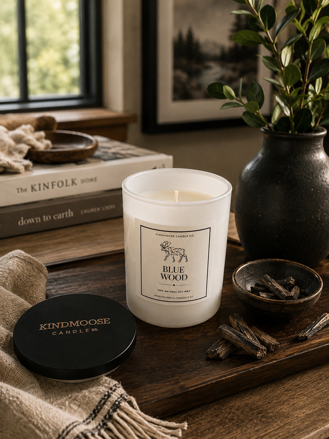 Blue Wood 6 oz soy candle by KINDMOOSE Candle Co. with the lid removed, styled on a rich wooden tray alongside charred wood pieces, a black ceramic vase with greenery, stacked books, and natural linen accents. Hand-poured in Canada and photographed in warm natural light to showcase its clean-burning soy wax and wood-inspired fragrance.