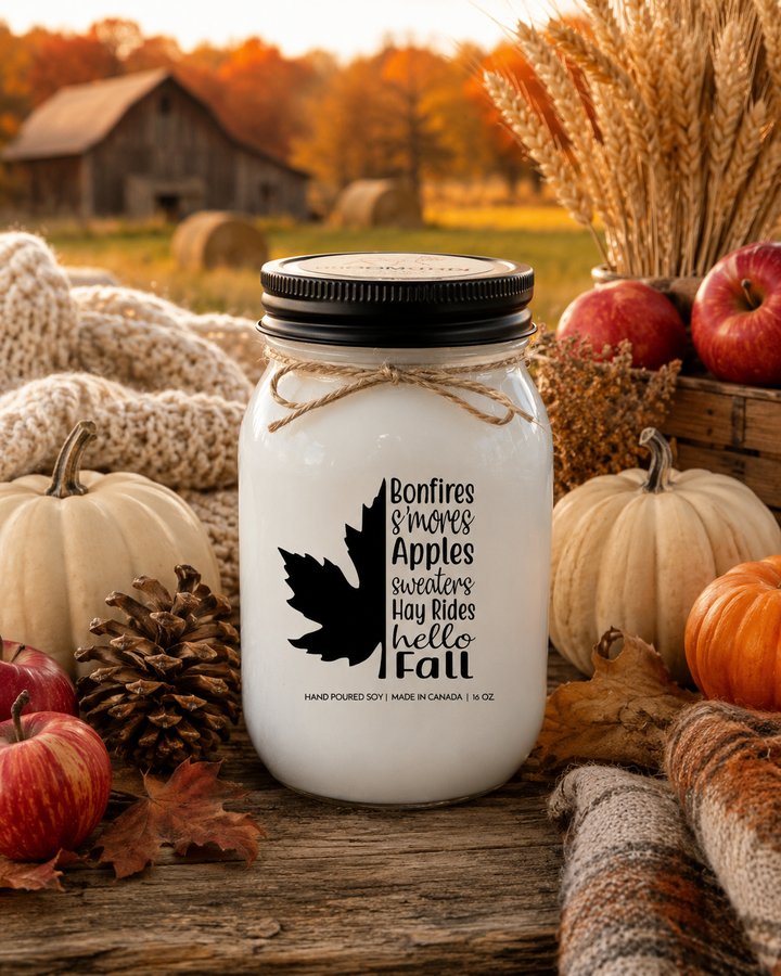 BONFIRES, S'MORES, APPLES, SWEATERS, HAY RIDES, HELLO FALL soy candle displayed on a rustic harvest table surrounded by apples, pumpkins, pinecones, autumn leaves, and countryside fall scenery.