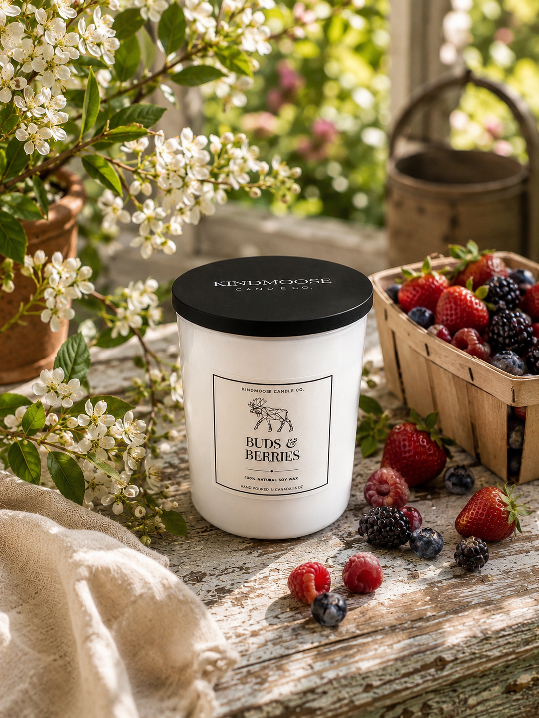 Buds & Berries 6 oz soy candle by KINDMOOSE Candle Co. displayed with fresh mixed berries, delicate spring blossoms, and natural greenery in a bright, premium cottage-inspired setting. Hand-poured in Canada with a black lid and clean minimalist label.