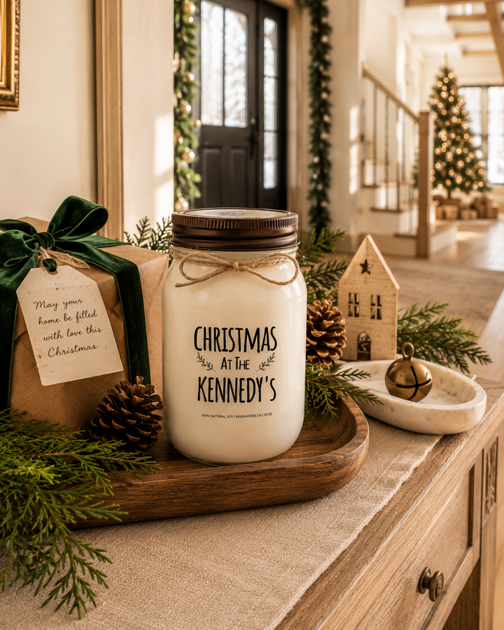 Personalized "Christmas at the..." soy candle with a distressed bronze lid displayed as a thoughtful Christmas gift on a farmhouse entry console with wrapped presents, cedar branches, pinecones, and elegant holiday décor.