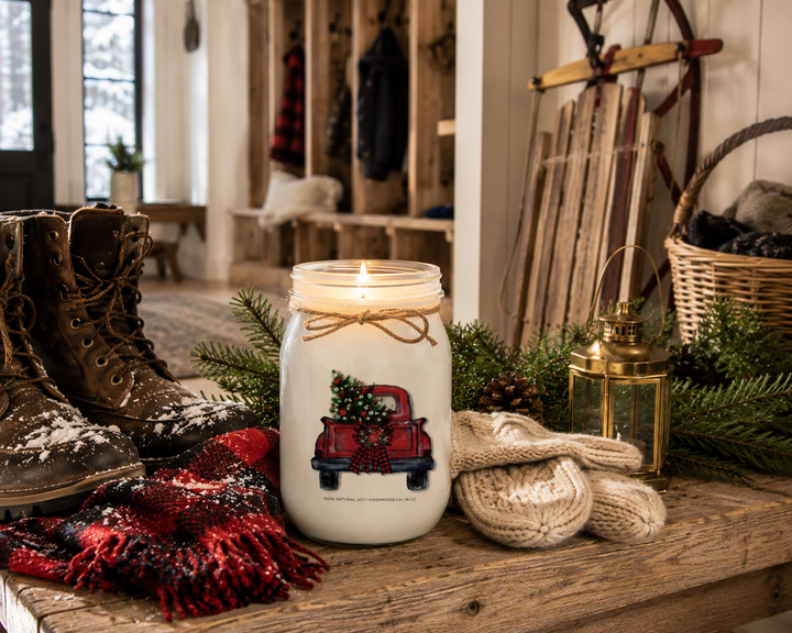 CHRISTMAS TREE TRUCK personalized Christmas candle burning on a reclaimed white oak mudroom bench beside snow-dusted winter boots, knit mittens, cedar branches, pinecones, and a vintage wooden sled in a cozy luxury farmhouse.