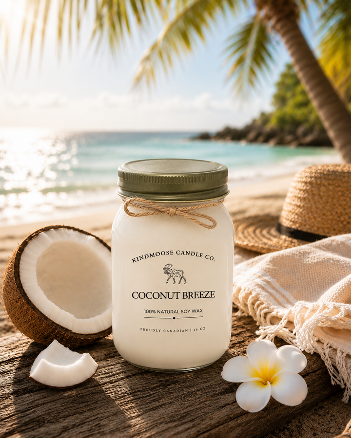 Coconut Breeze soy candle by KINDMOOSE Candle Co. displayed on weathered driftwood along a sunlit tropical beach. A frosted white mason jar candle with a bronze lid is surrounded by a fresh coconut, white plumeria flower, woven beach hat, and soft white towel. Palm trees frame the background while turquoise ocean waves and warm golden sunlight create a relaxing coastal atmosphere.