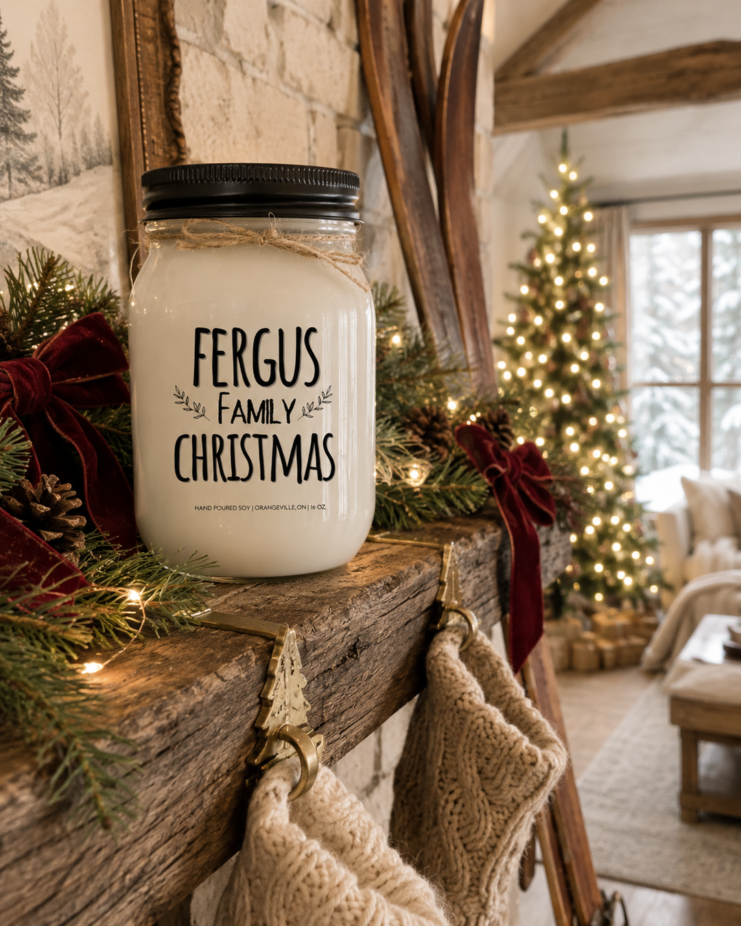 Personalized FAMILY CHRISTMAS candle by KINDMOOSE displayed on a rustic reclaimed wood mantel with a black lid, frosted white mason jar, and twine bow. Fresh evergreen garland, soft white Christmas lights, red velvet bows, pinecones, brass stocking holders, cream knit stockings, and a beautifully decorated farmhouse Christmas tree create a warm and inviting holiday living room filled with timeless family traditions.