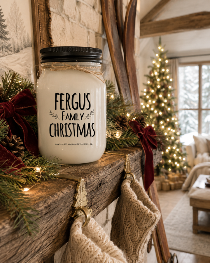 Personalized FAMILY CHRISTMAS candle by KINDMOOSE displayed on a rustic reclaimed wood mantel with a black lid, frosted white mason jar, and twine bow. Fresh evergreen garland, soft white Christmas lights, red velvet bows, pinecones, brass stocking holders, cream knit stockings, and a beautifully decorated farmhouse Christmas tree create a warm and inviting holiday living room filled with timeless family traditions.