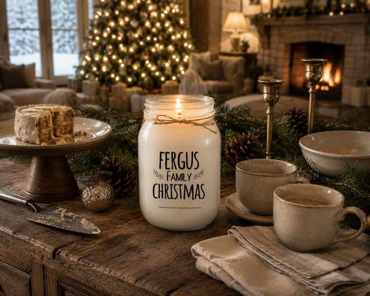 Personalized FAMILY CHRISTMAS candle by KINDMOOSE burning softly without a lid on a rustic reclaimed wood sideboard after Christmas dinner. The frosted white mason jar and twine bow remain unchanged while a slice of Christmas cake, coffee cups, evergreen garland, pinecones, vintage ornaments, and a softly glowing Christmas tree create a peaceful evening filled with gratitude, cherished memories, and the warmth of family traditions.
