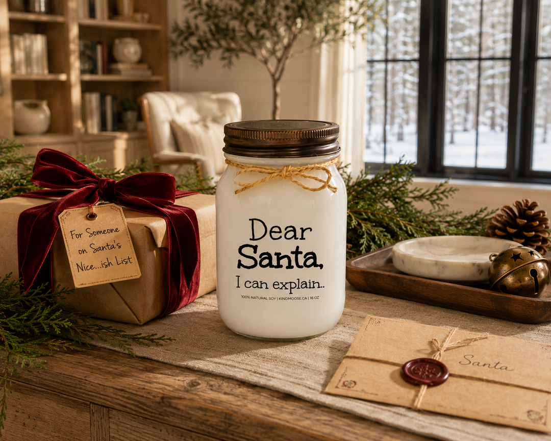 KINDMOOSE Dear Santa, I Can Explain candle with a distressed bronze lid displayed on a reclaimed white oak library console beside a beautifully wrapped Christmas gift with cranberry velvet ribbon, a handwritten gift tag, cedar branches, pinecones, a vintage letter to Santa, and elegant holiday décor in a luxury Canadian modern farmhouse.