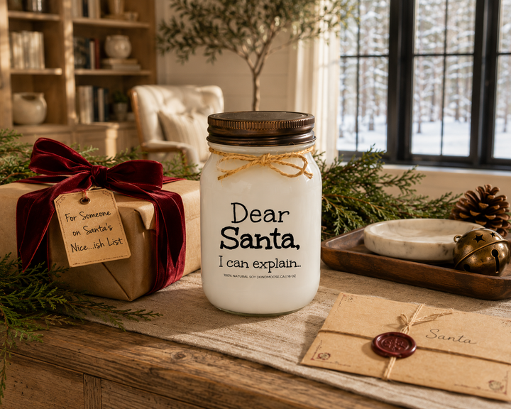 KINDMOOSE Dear Santa, I Can Explain candle with a distressed bronze lid displayed on a reclaimed white oak library console beside a beautifully wrapped Christmas gift with cranberry velvet ribbon, a handwritten gift tag, cedar branches, pinecones, a vintage letter to Santa, and elegant holiday décor in a luxury Canadian modern farmhouse.