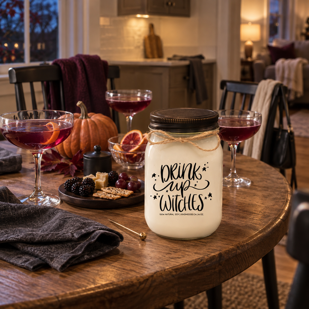 DRINK UP WITCHES KINDMOOSE candle with distressed bronze lid on an oak table during a cozy Halloween girls' night with friends.