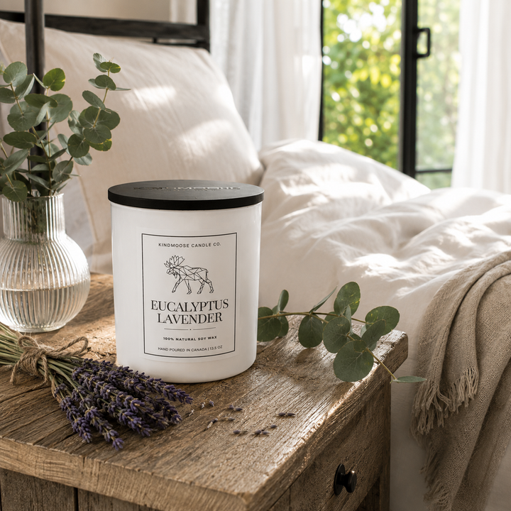 Eucalyptus Lavender Double Wick soy candle in a straight-sided painted white vessel with a black wooden lid, displayed on a reclaimed wood bedside table beside fresh eucalyptus and lavender in a bright, sunlit bedroom.