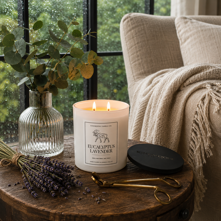 Burning Eucalyptus Lavender Double Wick soy candle with two flames glowing beside fresh eucalyptus, lavender and an antique wick trimmer on a reclaimed wood table overlooking rain-covered windows in a peaceful reading nook.