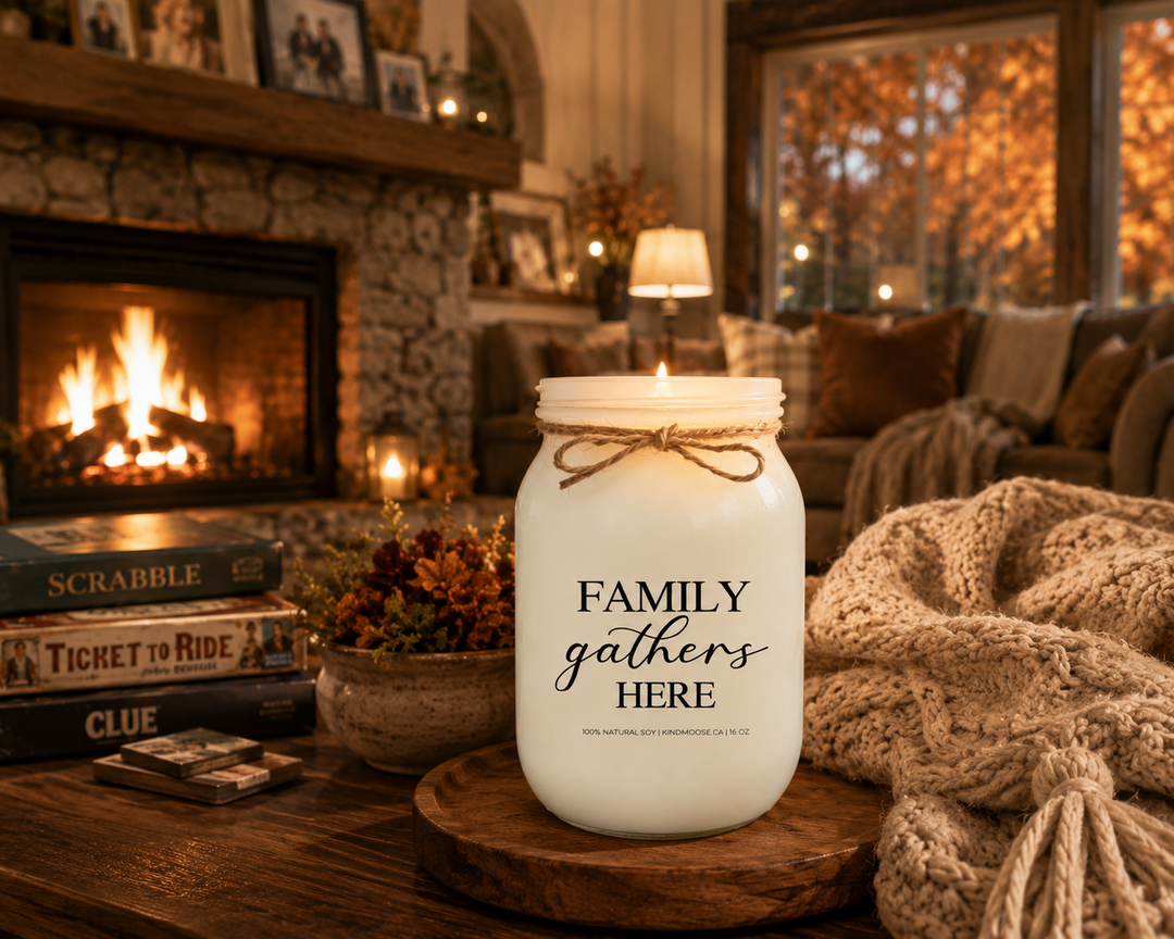 Burning FAMILY GATHERS HERE soy candle in a cozy farmhouse living room featuring a natural stone fireplace, family photographs, autumn greenery, soft knit blankets, and warm evening light. The scene celebrates home, belonging, and the comfort of family traditions.
