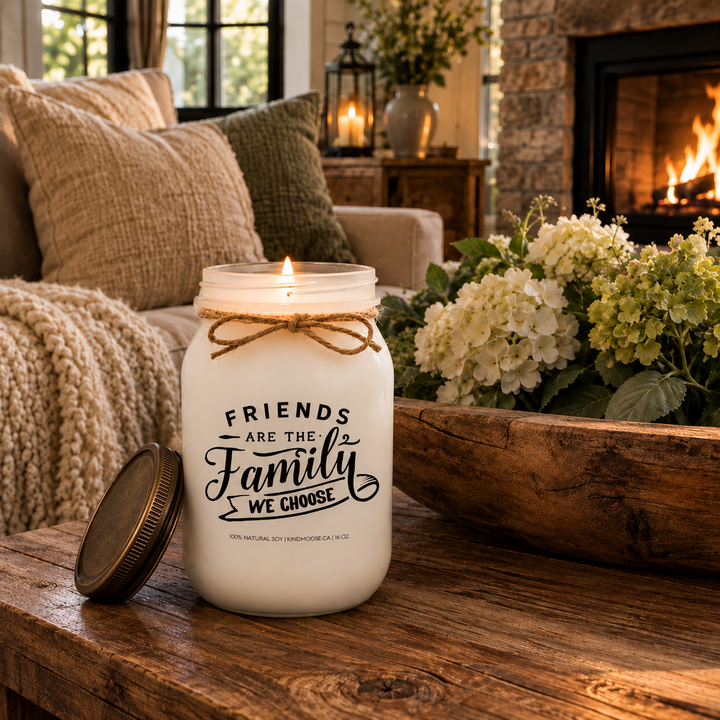 KINDMOOSE Friends Are The Family We Choose frosted soy candle burning on a cozy farmhouse coffee table beside a fireplace in a warm living room.