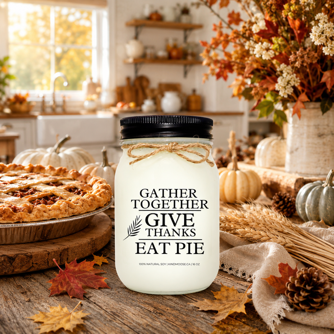 GATHER TOGETHER • GIVE THANKS • EAT PIE soy candle displayed on a rustic farmhouse harvest table beside a freshly baked pie, mini pumpkins, wheat stems, pinecones, and autumn leaves. Warm sunlight fills a cozy farmhouse kitchen, creating a welcoming atmosphere that celebrates gratitude, family traditions, and the comforts of fall.