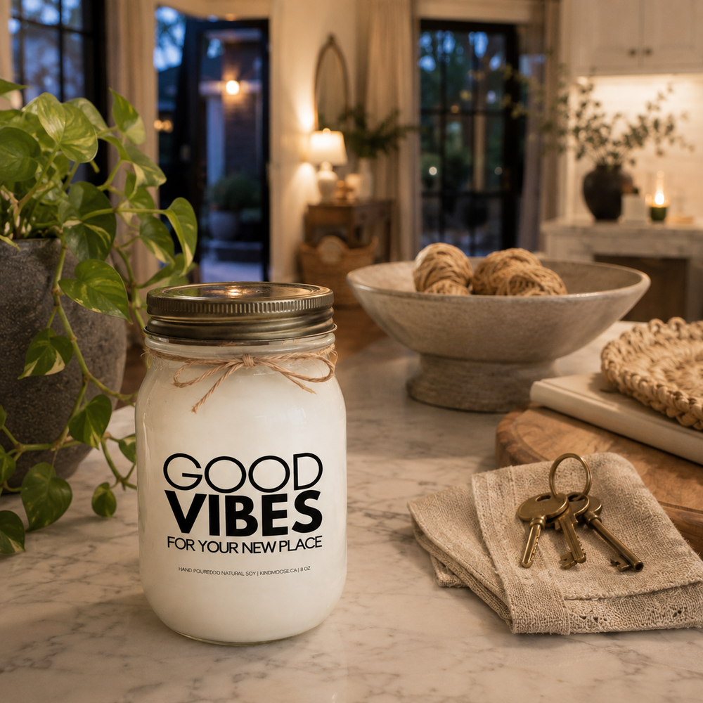 KINDMOOSE Good Vibes For Your New Place frosted soy candle on a white oak coffee table in a newly furnished living room with house plans and new home keys.