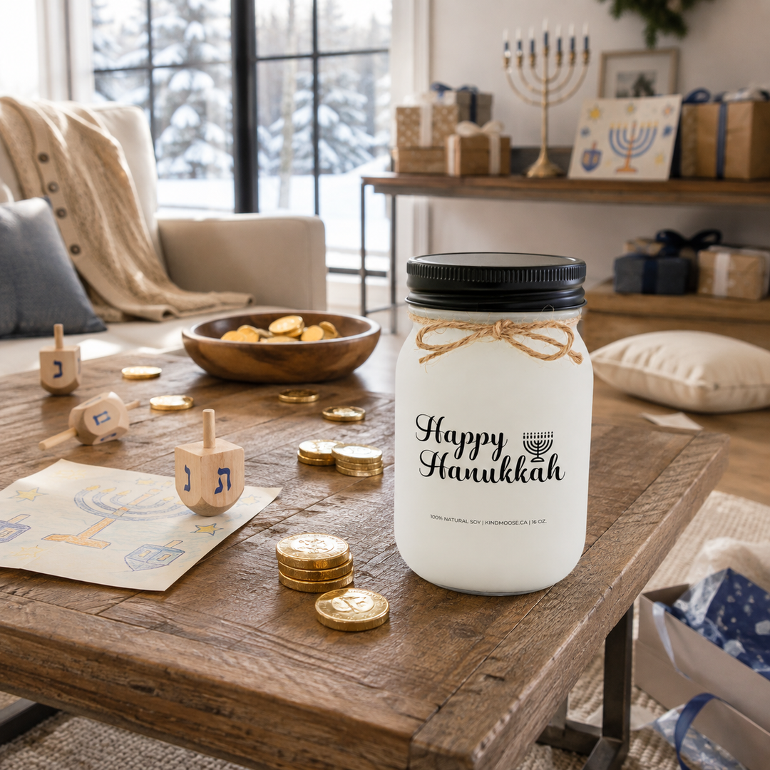 KINDMOOSE Happy Hanukkah 16 oz candle with black lid beside wooden dreidels and chocolate gelt during a family Hanukkah game.