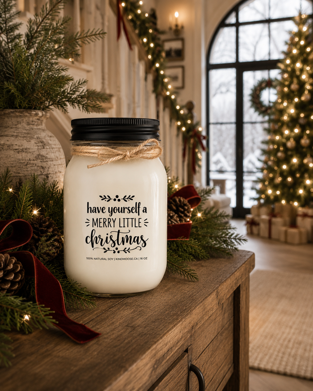 Have Yourself a Merry Little Christmas KINDMOOSE soy candle with a black lid displayed on a rustic white oak fireplace mantel decorated with fresh cedar garland, cream knit stockings, pinecones, warm white Christmas lights, and deep red velvet ribbon inside a cozy luxury farmhouse living room.