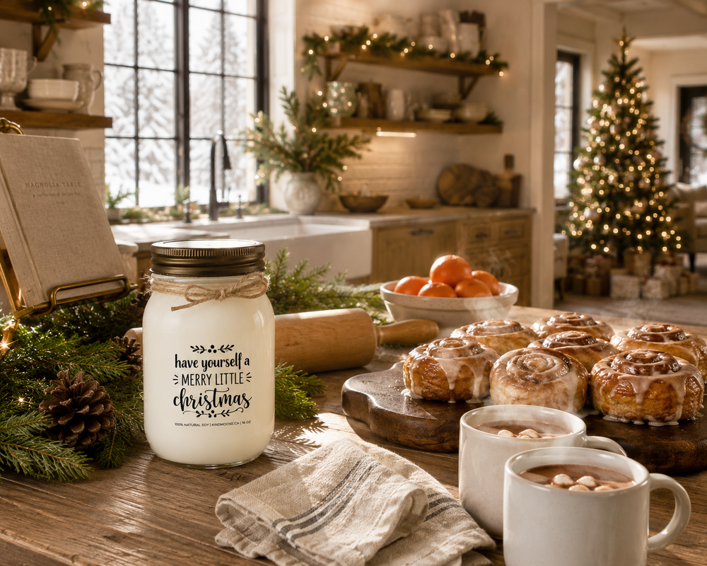 Have Yourself a Merry Little Christmas KINDMOOSE soy candle with a distressed bronze lid on a reclaimed white oak kitchen island surrounded by freshly baked cinnamon rolls, steaming hot chocolate, clementines, cedar sprigs, and holiday baking accents in a bright luxury farmhouse kitchen.