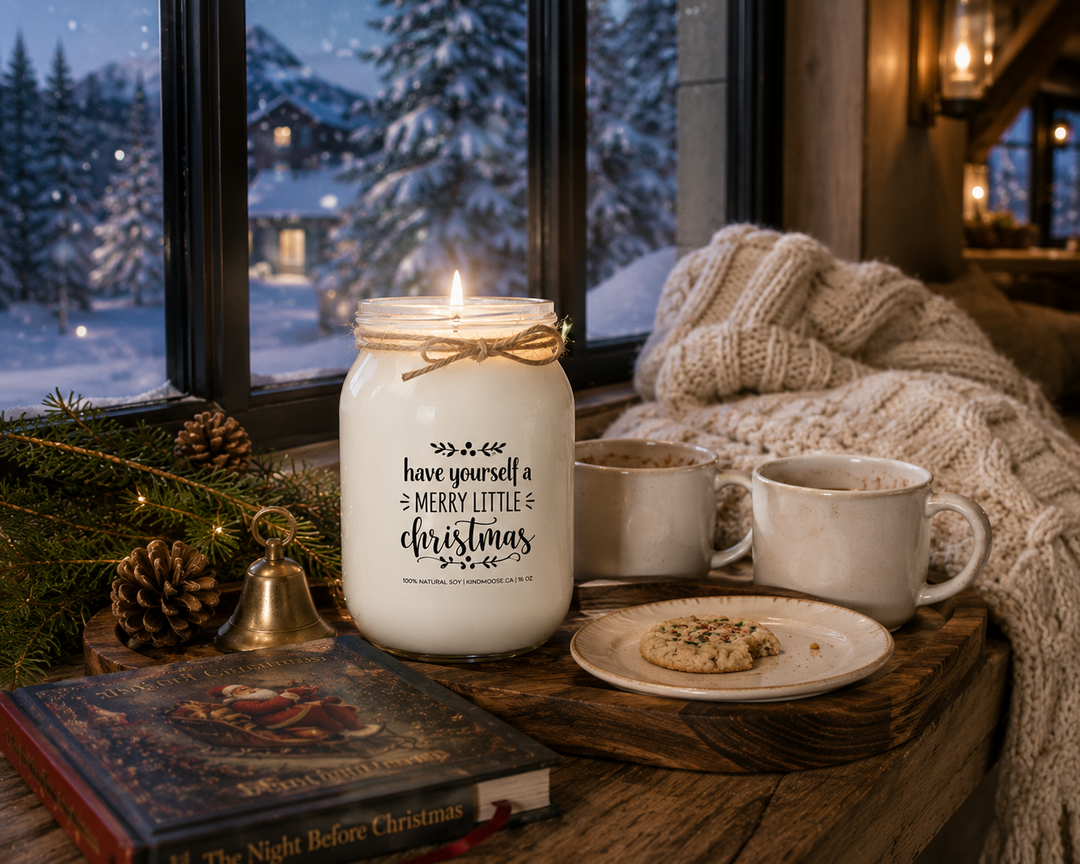 Lit Have Yourself a Merry Little Christmas KINDMOOSE soy candle on a reclaimed walnut window bench overlooking a snowy winter landscape, styled with a cream knit throw, Christmas storybook, hot chocolate mugs, a holiday cookie, cedar sprigs, pinecones, and a vintage brass bell for a peaceful Christmas evening.
