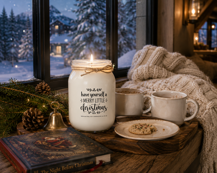 Lit Have Yourself a Merry Little Christmas KINDMOOSE soy candle on a reclaimed walnut window bench overlooking a snowy winter landscape, styled with a cream knit throw, Christmas storybook, hot chocolate mugs, a holiday cookie, cedar sprigs, pinecones, and a vintage brass bell for a peaceful Christmas evening.