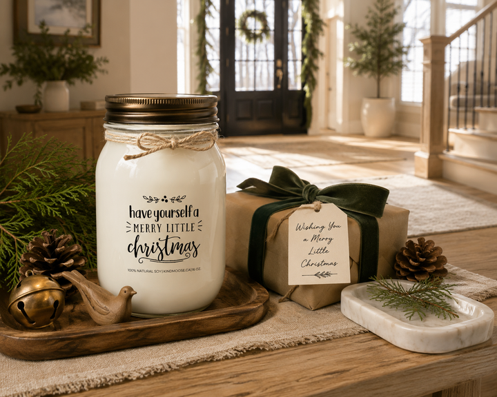 Have Yourself a Merry Little Christmas KINDMOOSE soy candle with a distressed bronze lid displayed on a reclaimed white oak entry console beside a beautifully wrapped Christmas gift with a forest green velvet ribbon, handwritten gift tag, cedar branches, pinecones, antique brass sleigh bell, and carved wooden dove ornament in a luxury farmhouse foyer.
