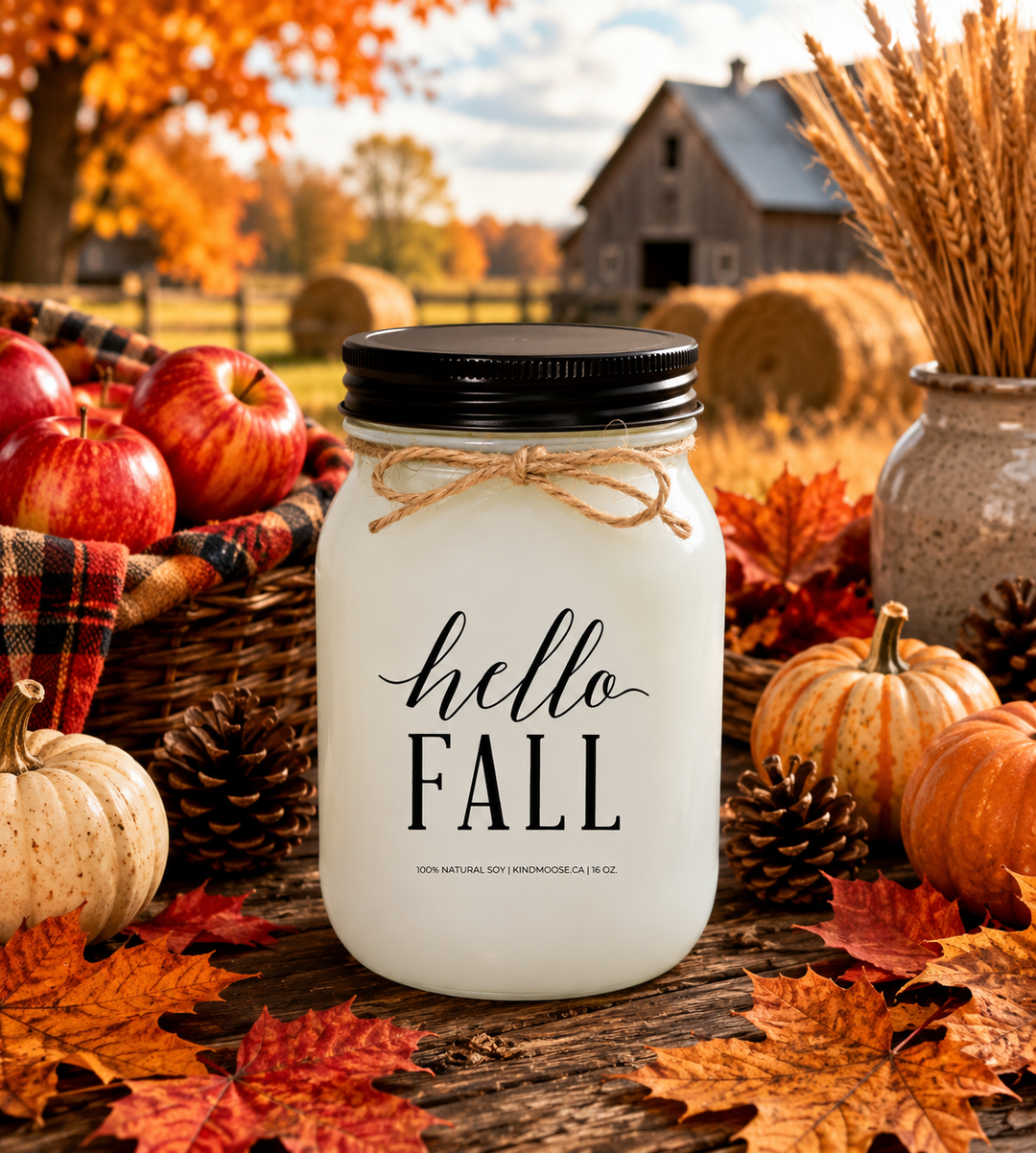 HELLO FALL soy candle displayed on a rustic farmhouse harvest table surrounded by colourful autumn leaves, red apples, mini pumpkins, pinecones, wheat stems, and cozy plaid accents. Warm countryside harvest scenery and golden fall sunlight create a welcoming atmosphere that celebrates the arrival of autumn.