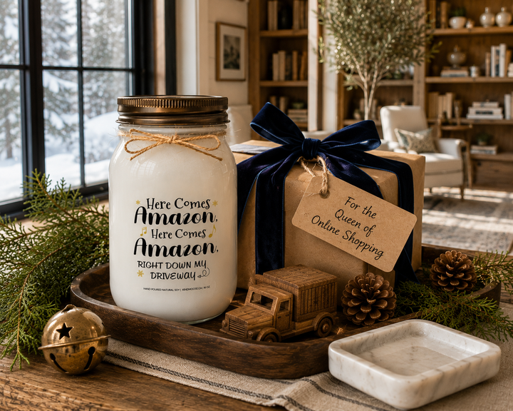 HERE COMES AMAZON, HERE COMES AMAZON, RIGHT DOWN MY DRIVEWAY candle presented as a humorous Christmas gift on a reclaimed white oak library console with a wrapped kraft paper present, navy velvet ribbon, cedar branches, pinecones, a carved wooden delivery truck ornament, and softly falling snow outside a luxury Canadian farmhouse library.