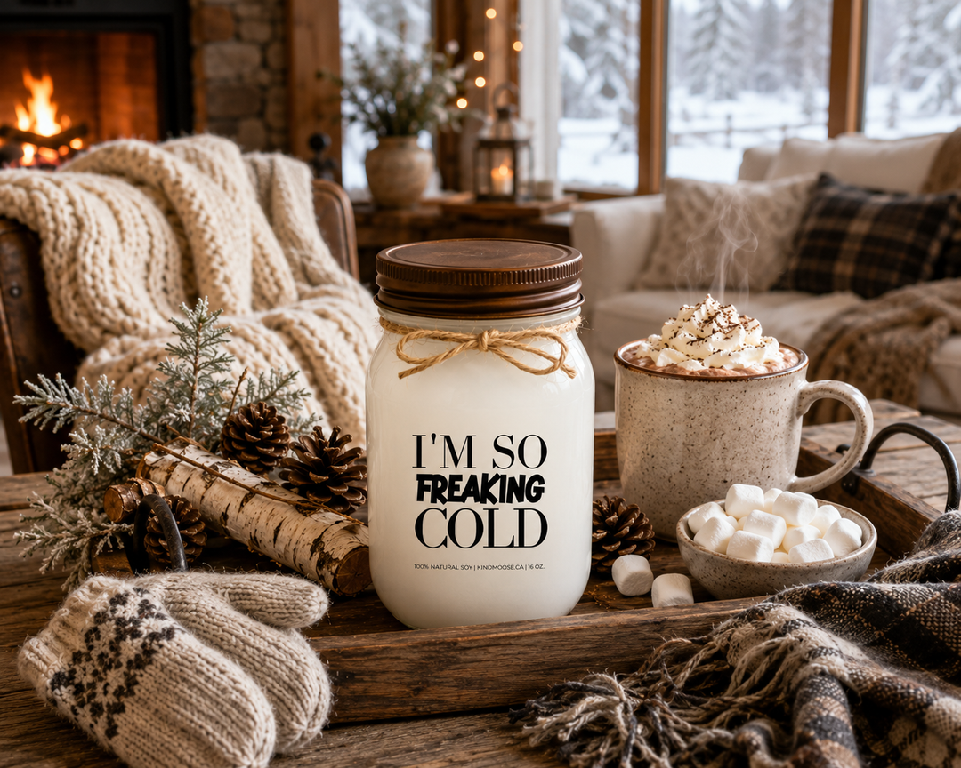 I’M SO FREAKING COLD soy candle with distressed bronze lid styled on a farmhouse table beside hot chocolate, marshmallows, wool mittens, knit blankets, pinecones, and winter greenery. The cozy winter gathering scene captures the feeling of warming up indoors after spending time in the cold.