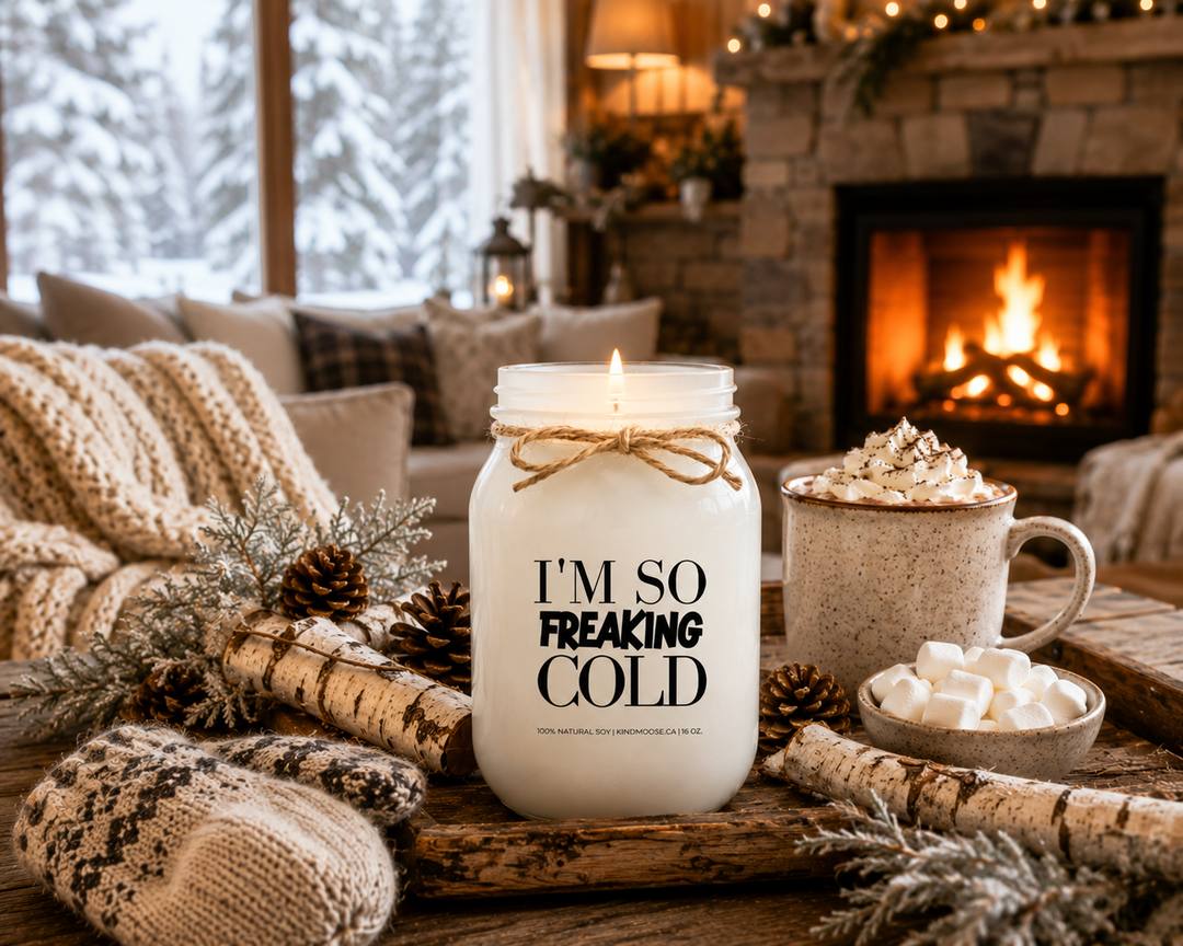 Burning I’M SO FREAKING COLD soy candle displayed in a cozy farmhouse living room featuring a stone fireplace, hot chocolate, chunky knit blanket, wool mittens, pinecones, birch accents, and snow-covered views outside. The warm winter scene celebrates comfort, relaxation, and escaping freezing temperatures.